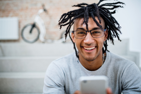 A portrait of young mixed race businessman with dreadlocks and smartphone in office, looking at camera.