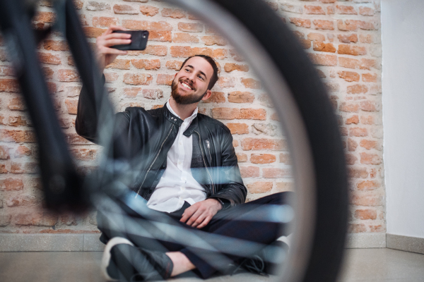 A young businessman with bicycle and telephone sitting on the floor in office, taking selfie.