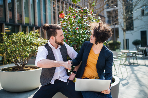 Young businesspeople with laptop outdoors in courtyard, shaking hands. Start-up concept.