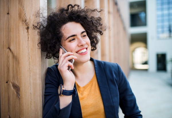 A happy young business woman with smartphone standing outdoors, making a phone call. Start-up concept.