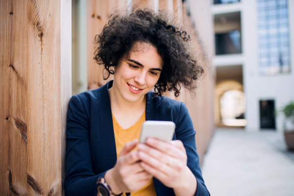 A happy young business woman with smartphone standing outdoors, text messaging. Start-up concept.