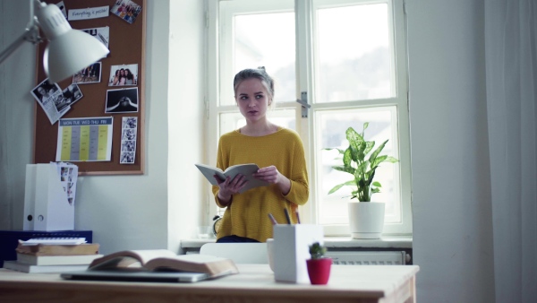 A young happy college female student with a book standing by window at home, studying.