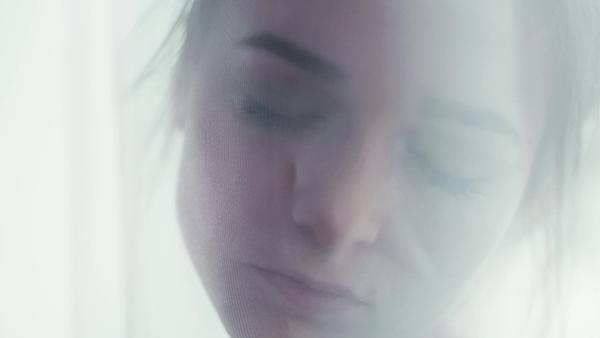 A portrait of young woman standing by window behind lace curtain, dreaming.