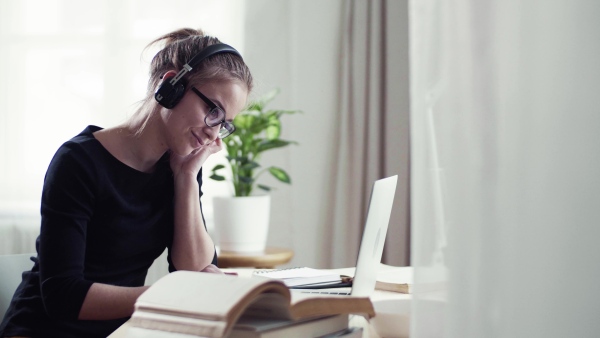 A young happy college female student sitting at the table at home, using headphones and laptop when studying.