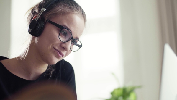 A young happy college female student sitting at the table at home, using headphone when studying.