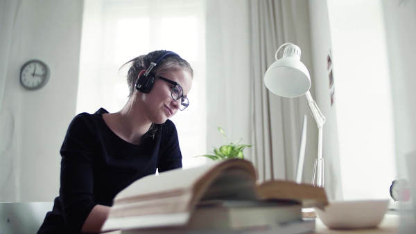 A young happy college female student sitting at the table at home, using headphones and laptop when studying.