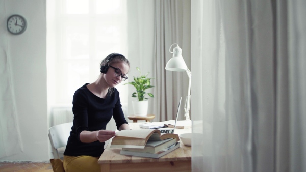 A young happy college female student sitting at the table at home, using headphones and tablet when studying.