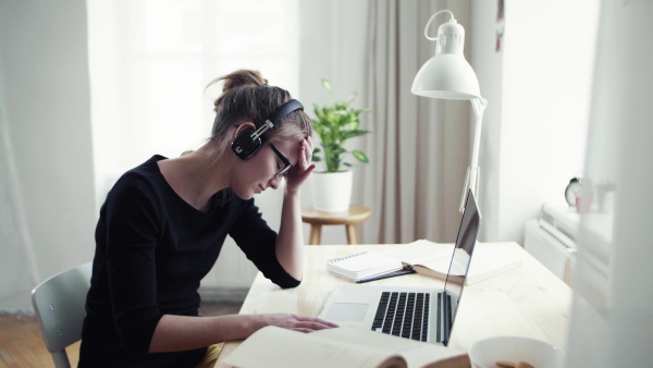 A young frustrated college female student sitting at the table at home, using headphones and laptop when studying.