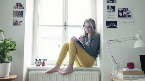 A young happy college female student with a book sitting on window sill at home, studying.