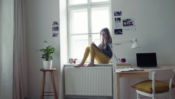 A young happy college female student with an exercise book sitting on window sill at home, studying.