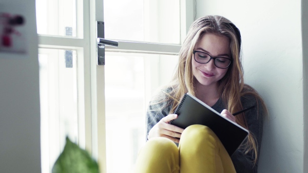 A young happy college female student with an exercise book sitting on window sill at home, studying.