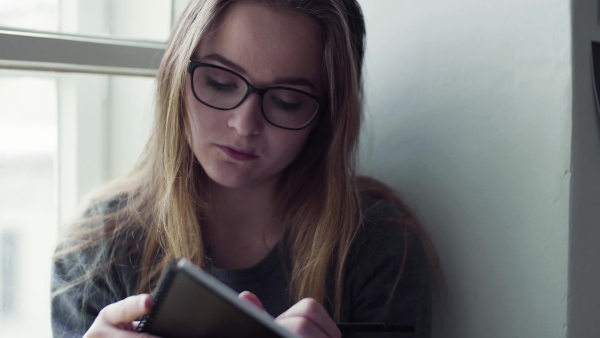 A young happy college female student with an exercise book sitting on window sill at home, writing.