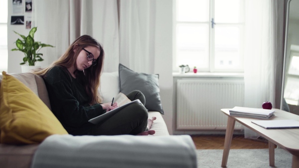 A happy young female student sitting on sofa, writing when studying.