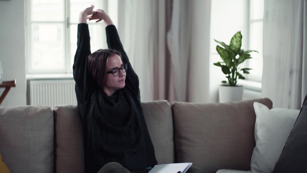 A tired young female student sitting on sofa, stretching when studying.