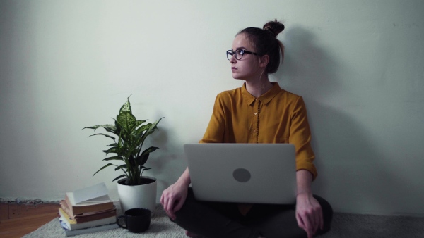 A sad young happy college female student with laptop sitting on floor at home, studying.