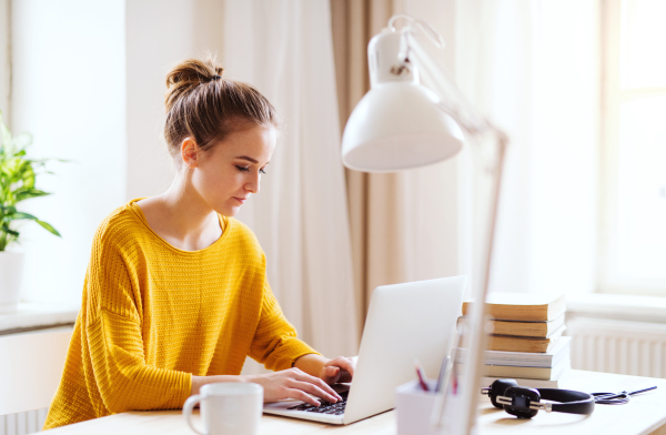 A young happy college female student sitting at the table at home, using laptop when studying.