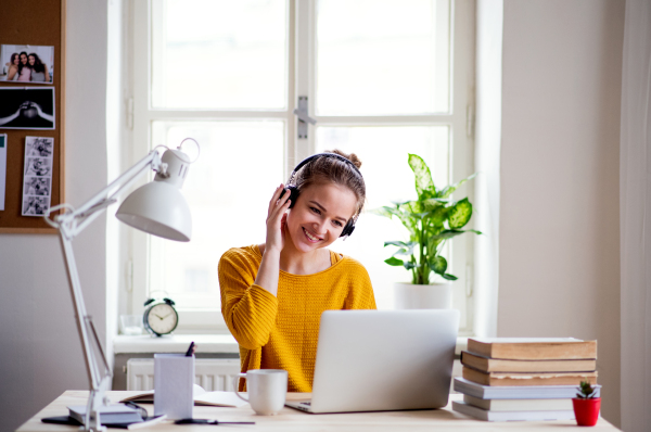 A young happy college female student sitting at the table at home, using laptop and headphones when studying.