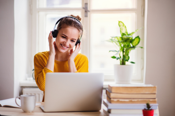 A young happy college female student sitting at the table at home, using laptop and headphones when studying.