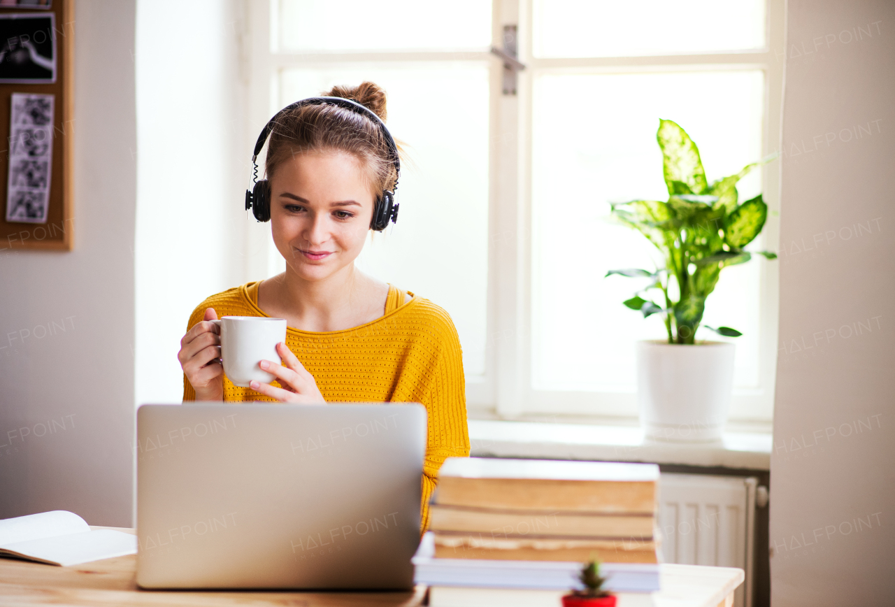 A young happy college female student sitting at the table at home, using headphones when studying.