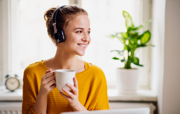 A young happy college female student sitting at the table at home, using headphones when studying.