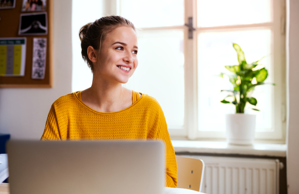 A young happy college female student sitting at the table at home, using laptop when studying.