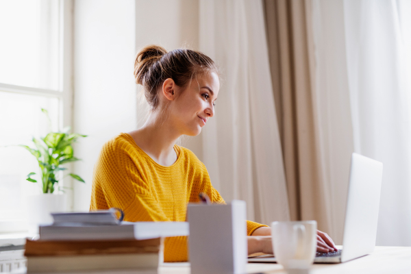 A young happy college female student sitting at the table at home, using laptop when studying.