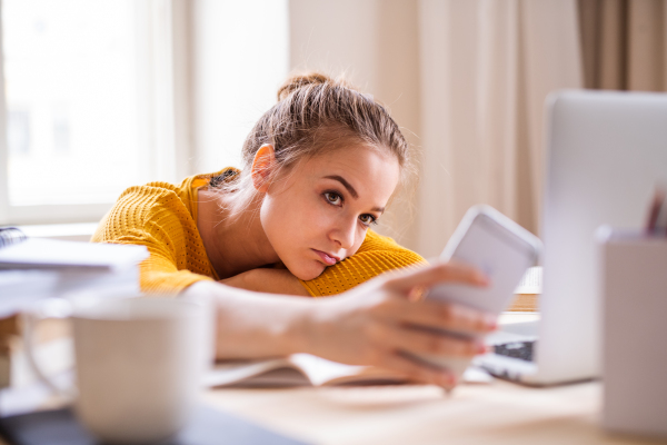 A young happy college female student sitting at the table at home, using laptop and smartphone when studying.