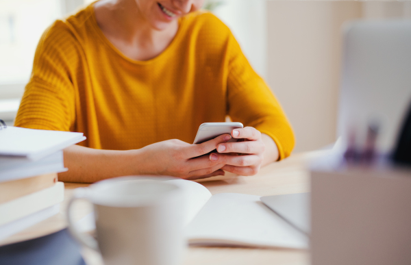 A midsection of young happy college female student sitting at the table at home, using smartphone when studying.