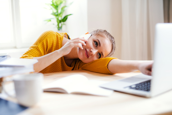 A young happy college female student sitting at the table at home, using laptop and smartphone when studying.