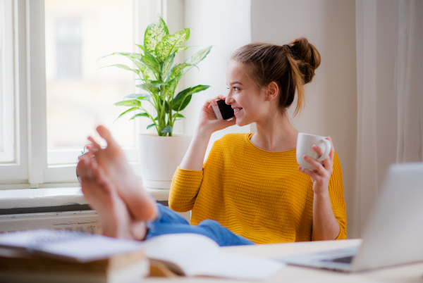 A young happy college female student sitting at the table at home, using smartphone when studying.