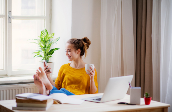 A young happy college female student sitting at the table at home, using smartphone when studying.