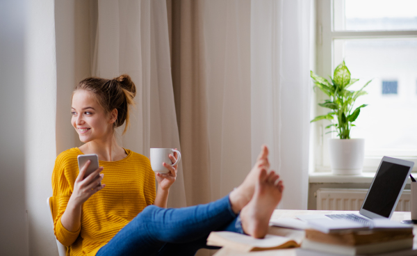 A young happy college female student sitting at the table at home, using smartphone when studying.