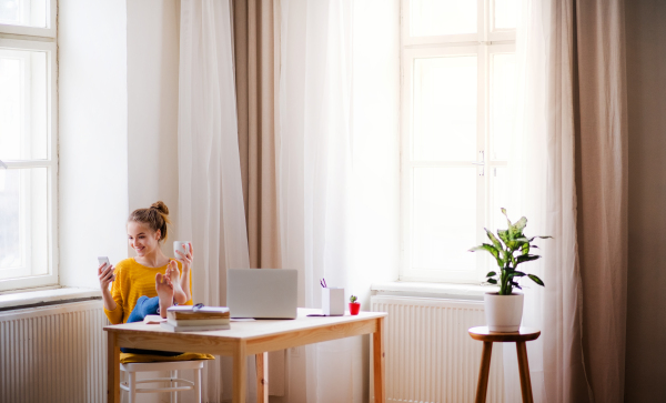 A young happy college female student sitting at the table at home, using smartphone when studying.