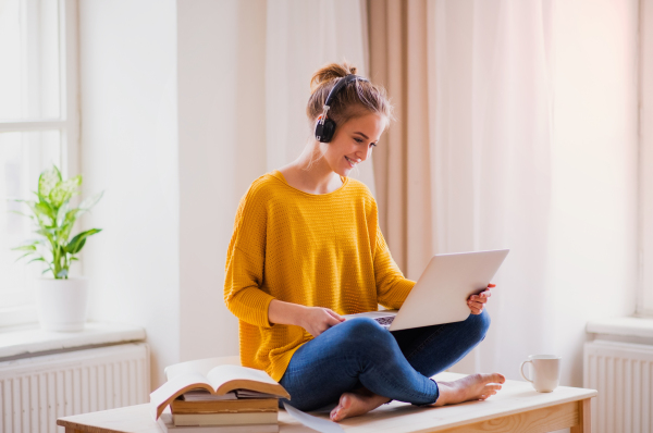 A young happy college female student sitting at the table at home, using headphones and laptop when studying.