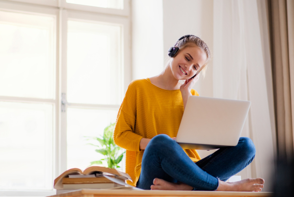 A young happy college female student sitting at the table at home, using headphones and laptop when studying.