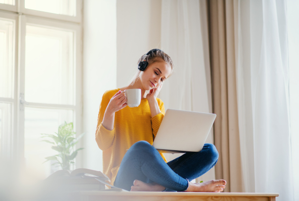 A young happy college female student sitting at the table at home, using headphones and laptop when studying.