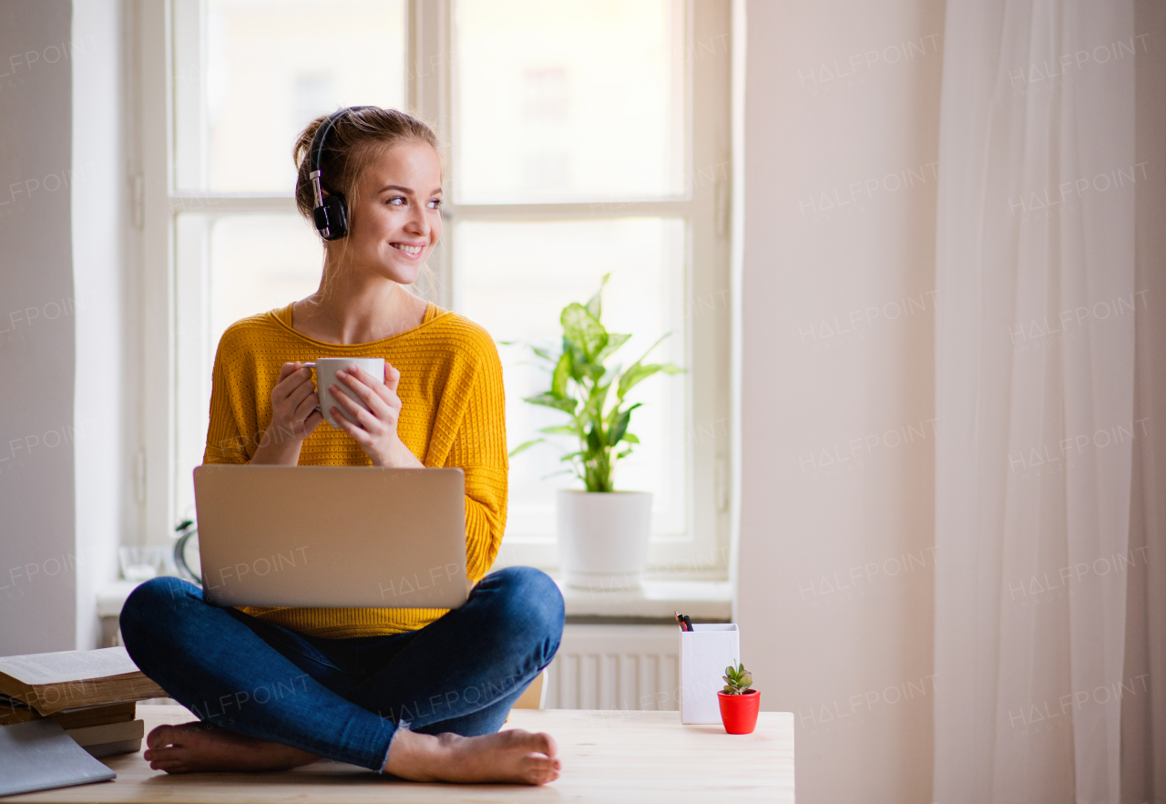 A young happy college female student sitting at the table at home, using headphones when studying.