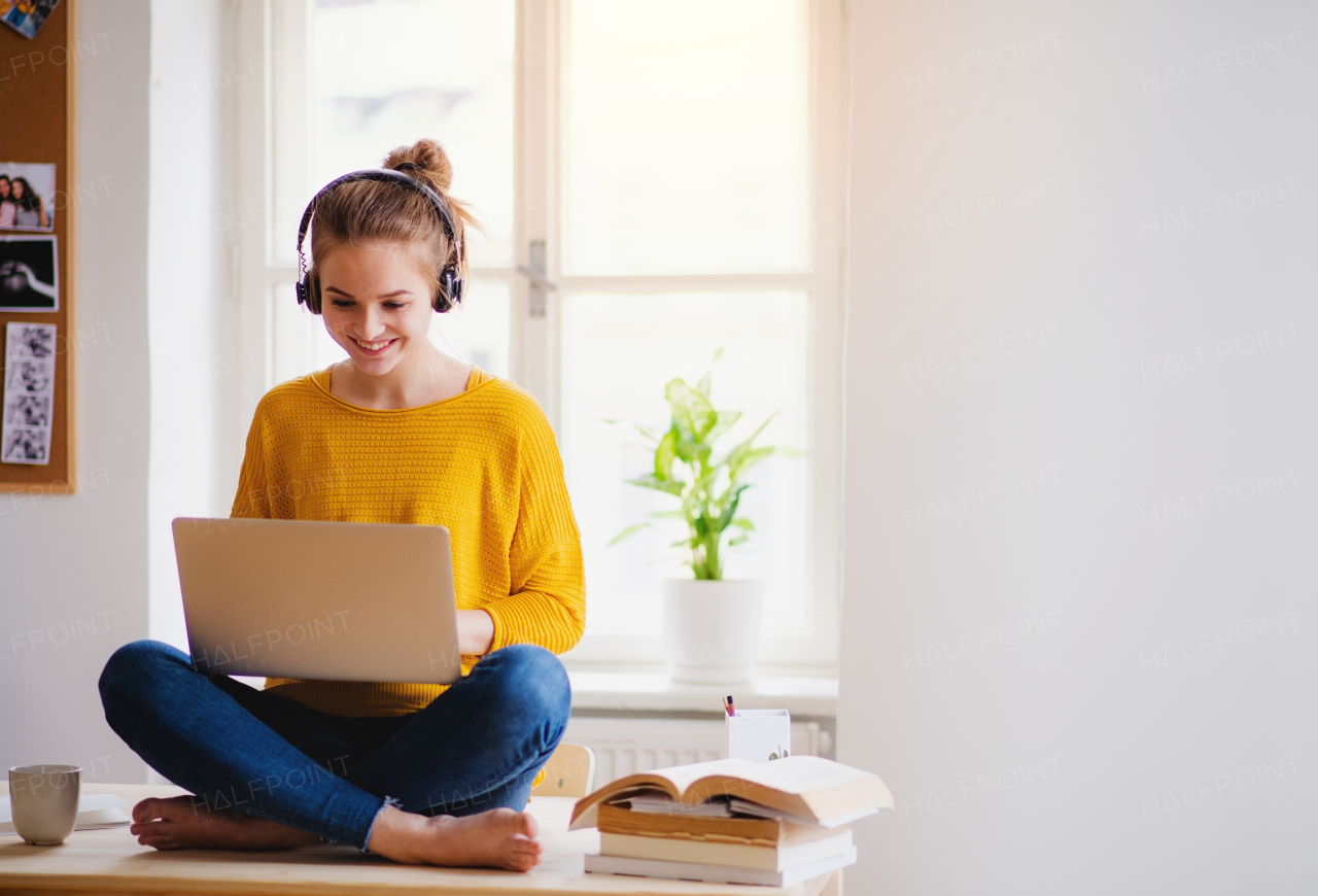 A young happy college female student sitting at the table at home, using headphones and laptop when studying.