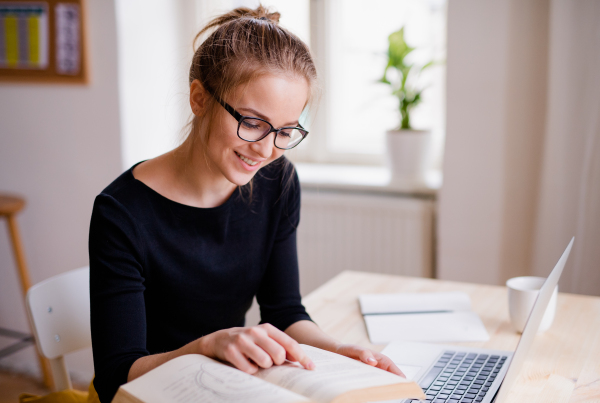 A young happy college female student sitting at the table at home, using laptop when studying.