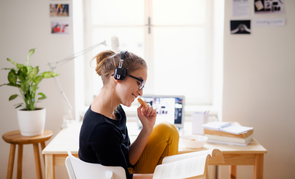 A young happy college female student sitting at the table at home, using headphones and laptop when studying.