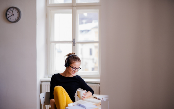 A young happy college female student sitting at the table at home, using headphones when studying.