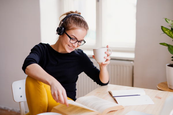 A young happy college female student sitting at the table at home, using headphones when studying.