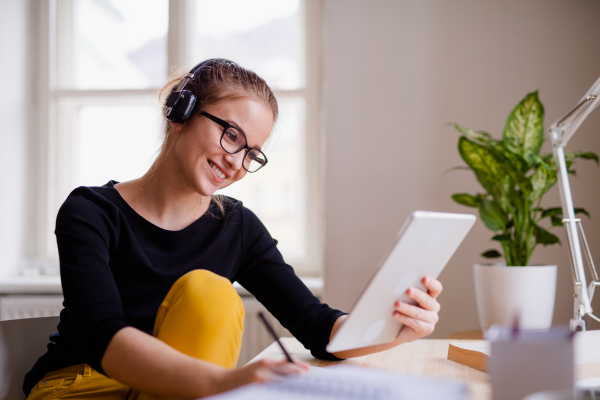 A young happy college female student sitting at the table at home, using headphones and tablet when studying.