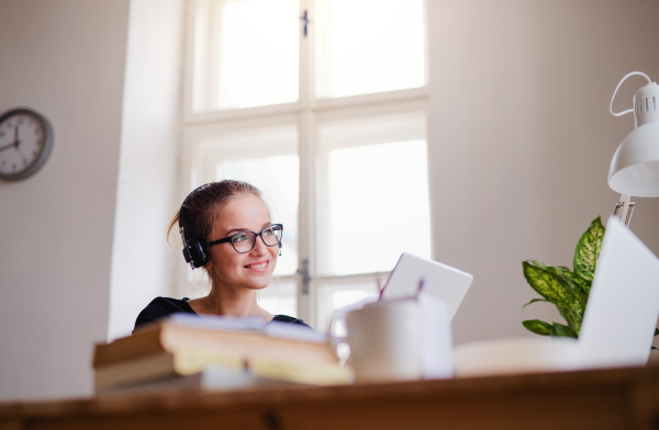 A young happy college female student sitting at the table at home, using headphones and tablet when studying.