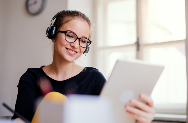 A young happy college female student sitting at the table at home, using headphones and tablet when studying.