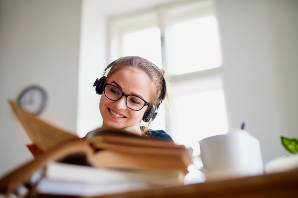 A young happy college female student sitting at the table at home, using headphones when studying.