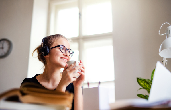 A young happy college female student sitting at the table at home, using headphones when studying.