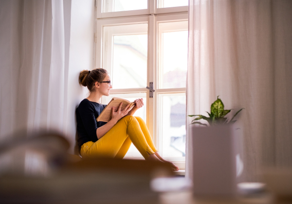 A young happy college female student with a book sitting on window sill at home, studying.