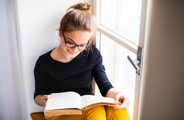 A young happy college female student with a book sitting on window sill at home, studying.