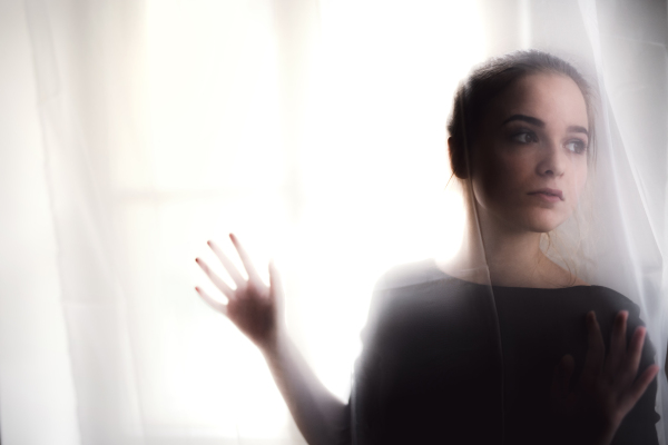 A silhouette of young sad woman standing by window. Copy space.